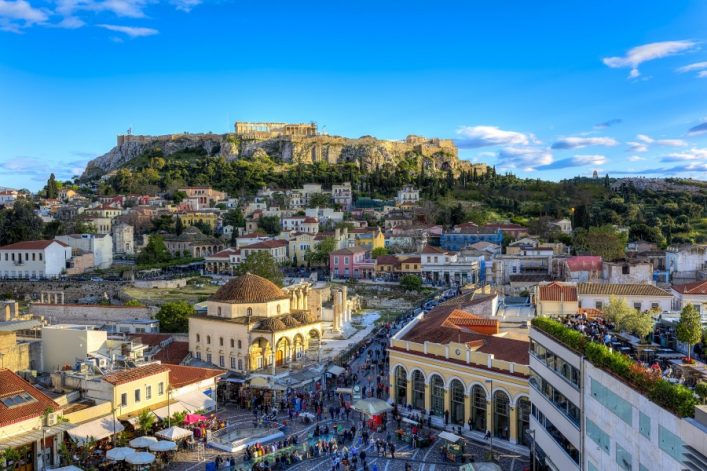 Blick auf die Akropolis in Athen, die majestätisch auf einem Hügel im Hintergrund thront. Im Vordergrund leuchtende Altstadtgebäude und eine belebte, von Menschen gefüllte Fußgängerzone bei klarem Himmel.
