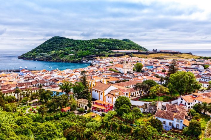 Blick auf eine Küstenstadt mit roten Dächern und weißen Gebäuden umgeben von üppigen Bäumen. Im Hintergrund erhebt sich ein grüner Hügel, der sich zum Meer hin öffnet, wo Boote auf dem Wasser liegen.