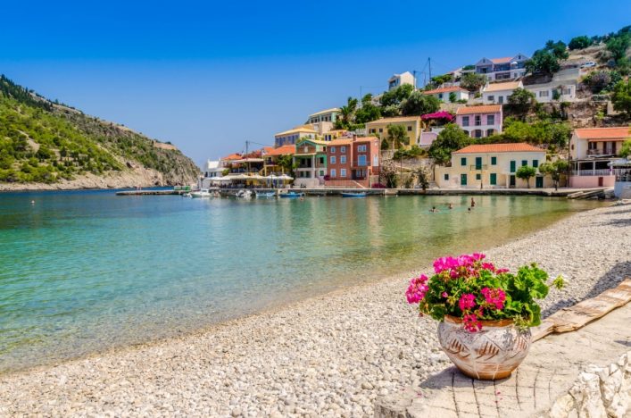 Strand mit Kieseln und einem Topf rosa Blumen im Vordergrund, blaues Meer links. Im Hintergrund bunte Häuser am Hang, türkisfarbenes Wasser mit badenden Menschen, grüne Hügel am Horizont.