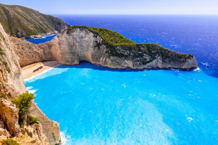 Hellblaues Meer umgibt die Bucht Navagio, umgeben von steilen Klippen mit grünem Bewuchs. Unten links ein Schiffswrack auf hellem Sandstrand. Im Hintergrund der tiefblaue Ozean.