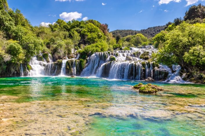 Ein breiter Wasserfall stürzt über mehrere Stufen in einen klaren, türkisfarbenen Fluss. Üppige grüne Bäume und Sträucher umgeben das Wasser, darüber ein blauer Himmel mit wenigen Wolken.