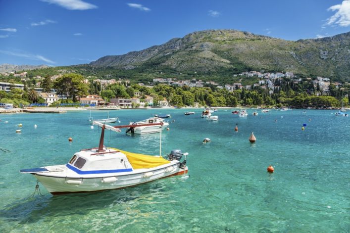 Klares türkisblaues Wasser, im Vordergrund ein kleines Boot mit gelbem Sonnenschutz. Im Hintergrund eine Küstenstadt mit weißen Häusern und grünen Hügeln unter blauem Himmel.