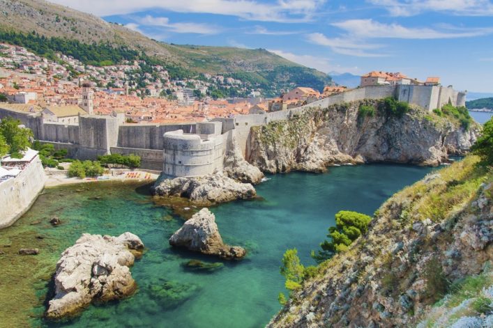 Blick auf Dubrovnik mit türkisem Meer, imposanten Stadtmauern und orangefarbenen Dächern im Hintergrund. Felsige Küste und grüne Vegetation im Vordergrund. Klare, blaue Himmel umrahmt die Szene.