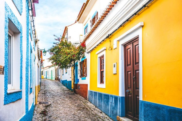 Pflasterstraße führt durch malerische Gasse in Ferragudo, Algarve, gesäumt von bunten Häusern: links blau-weiß, rechts gelb-orange mit braunen Türen. Ein Baum mit roten Blumen ragt hervor.