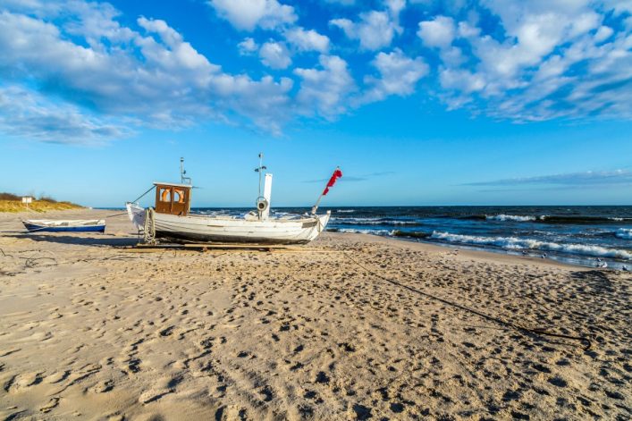 Ein weißes Fischerboot mit roter Flagge liegt auf einem sandigen Strand unter blauem Himmel, das Meer im Hintergrund. Links ist ein weiteres kleines Boot. Dünengras wächst links am Ufer.
