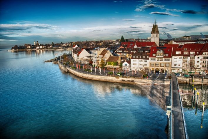 Die Uferpromenade von Friedrichshafen am Bodensee, gesäumt von weißen und gelben Gebäude, mit einer markanten Kirche im Hintergrund. Der blaue Himmel und das ruhige Wasser reflektieren die malerische Stadtansicht.