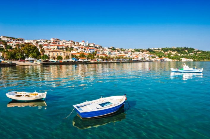 Blaues Wasser mit drei Booten im Vordergrund, dahinter eine Küstenstadt auf einem grünen Hügel, hellblaue und weiße Häuser unter klarem Himmel.