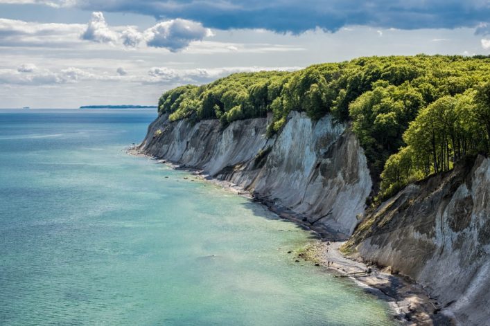 Steilküste mit weißen Felsen und grünen Bäumen am oberen Rand erstreckt sich von rechts unten nach links. Das Meer glitzert türkisblau unter wolkigem Himmel. Ein paar Menschen sind am Strand.