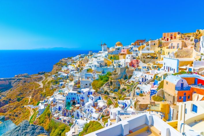 Weißgetünchte Häuser mit blauen Kuppeln und farbigen Akzenten stapeln sich malerisch auf einem Hügel in Oia, Santorin. Im Hintergrund leuchtet das blaue Meer unter einem klaren Himmel.