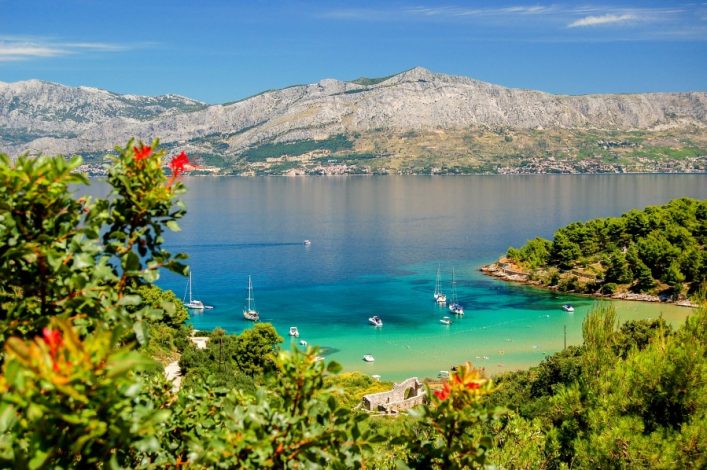 Klarer Himmel über Lovrečina Beach auf Brač, Kroatien: Im Vordergrund grüne Büsche mit roten Blüten, darunter türkisblaues Wasser mit Segelbooten, rechts dichter Wald, im Hintergrund Berge.