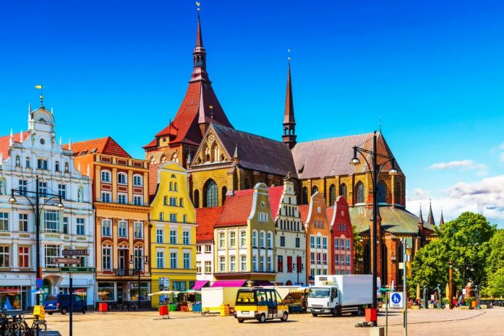 Bunte Giebelhäuser und eine große Kirche mit spitzen Türmen in Rostock, Deutschland. Die Gebäude ziehen sich links nach rechts, Bäume und ein blauer Himmel im Hintergrund. Autos und LKWs im Vordergrund.