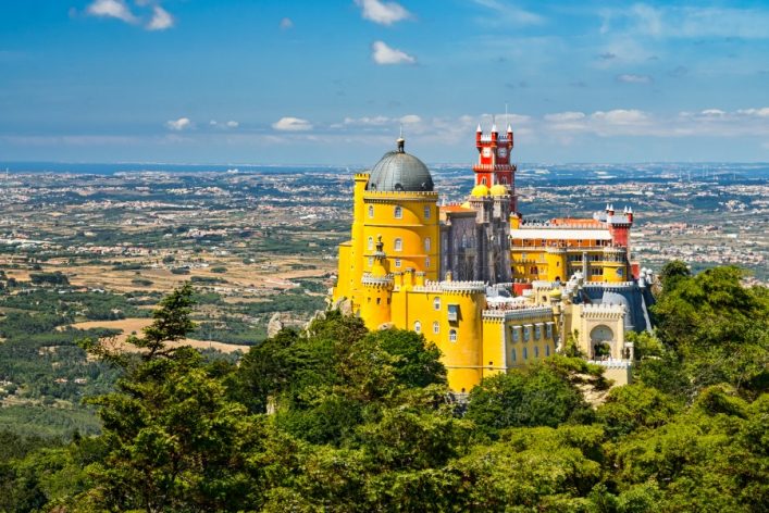 Bunte Burg mit gelben Fassaden und roten Türmen auf Hügel, umgeben von grünen Bäumen. Im Hintergrund weite Landschaft unter blauem Himmel mit wenigen Wolken.