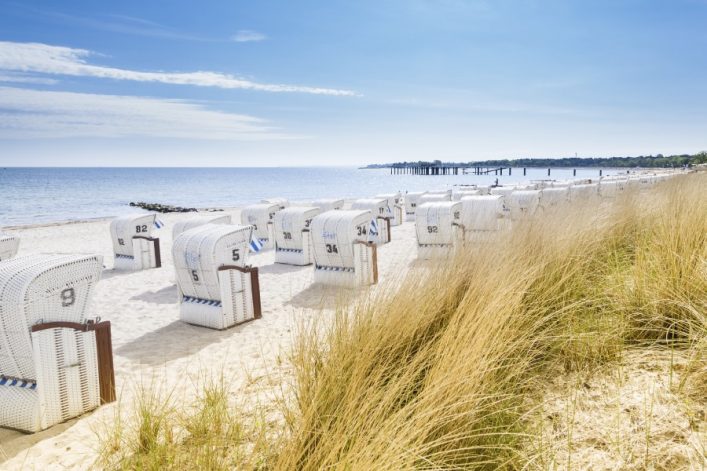 Sonne scheint auf einen Strand mit weißen Strandkörben in Reihen. Rechts im Vordergrund wachsen hohe Gräser, links erstreckt sich das blaue Meer bis zum Horizont. Am Strandhorizont ist ein Pier.