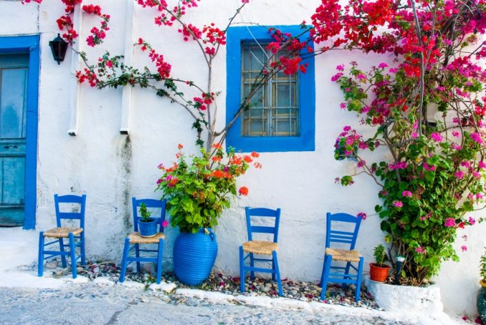 Vier blaue Holzstühle stehen links von einem Fenster mit blauen Rahmen in einer weißen Hausfassade auf der Insel Kos. Rote Bougainvillea rankt sich an der Wand, Pflanzen in Töpfen schmücken die Szene.