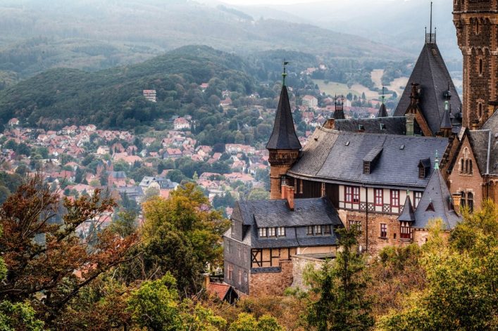 Wernigerode Schloss in der Mitte, graue Schieferdächer, umgeben von Bäumen. Im Hintergrund Häuser der Stadt am Hang, grüne Hügel erstrecken sich bis zum Horizont und bewölkter Himmel.