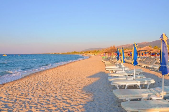 Sonniger Sandstrand mit azurblauem Meer links. Rechts stehen Reihen von weißen Liegestühlen und blauen Sonnenschirmen. Im Hintergrund sanfte Hügel und Palmen.