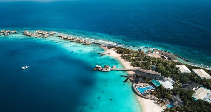 Ein tropisches Inselresort mit über Wasser gebauten Villen erstreckt sich entlang einer türkisfarbenen Lagune. Im Vordergrund sind Palmen und ein Pool zu sehen, rechts das offene Meer.
