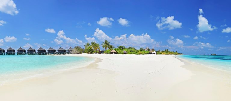 Strand mit weißem Sand und türkisfarbenem Wasser, im Hintergrund stehen auf dem Meer Holzhütten auf Stelzen, dahinter Palmen und üppige Vegetation. Der Himmel ist klar mit vereinzelten Wolken.