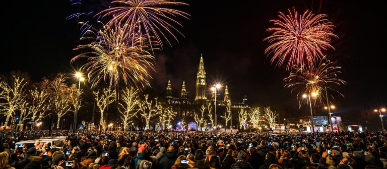 Eine große Menschenmenge sammelt sich unter buntem Feuerwerk am Nachthimmel vor dem Wiener Rathaus. Bäume mit Lichterketten stehen im Vordergrund, während viele mit ihren Handys fotografieren.
