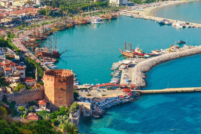 Roter Kizil Kule Turm in Alanya an der türkischen Riviera, umgeben von der historischen Festungsmauer, daneben blau schimmernder Hafen mit Booten, grün bewachsene Landschaft im Hintergrund.