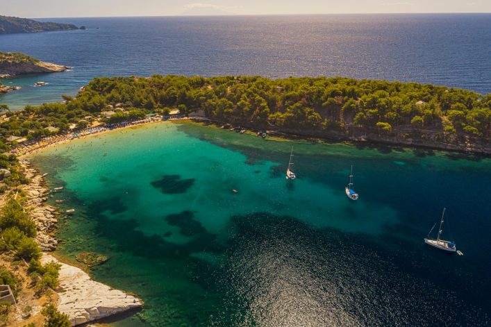 Luftaufnahme eines türkisfarbenen Strandes mit dicht bewaldeten Hügeln im Hintergrund. Drei Segelboote liegen auf ruhigem Wasser vor Anker. Links sind helle Felsformationen zu sehen.