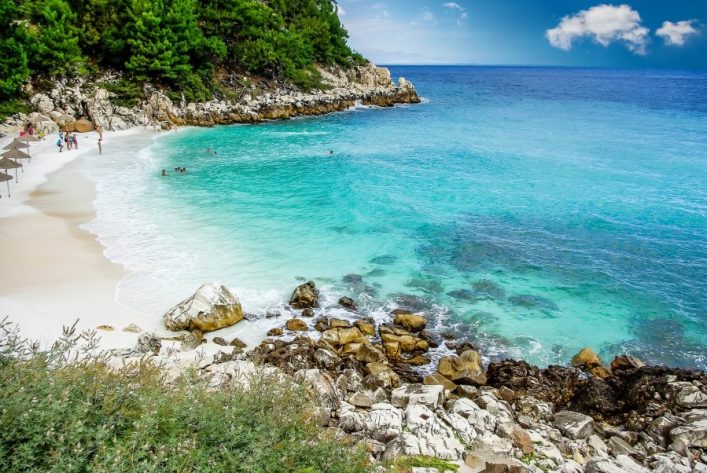 Azurblaues Meer trifft auf weißen Sandstrand, umgeben von üppigem Wald. Links sonnenbaden Menschen unter Schirmen, rechts liegen Felsen im klaren Wasser. Ein ruhiges, paradiesisches Szenario.