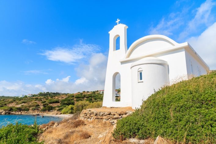 Weiße Kirche mit Glockenturm auf einem Hügel, umgeben von grünen Büschen. Im Hintergrund das blaue Meer und eine Hügelkette unter einem klaren, blauen Himmel.