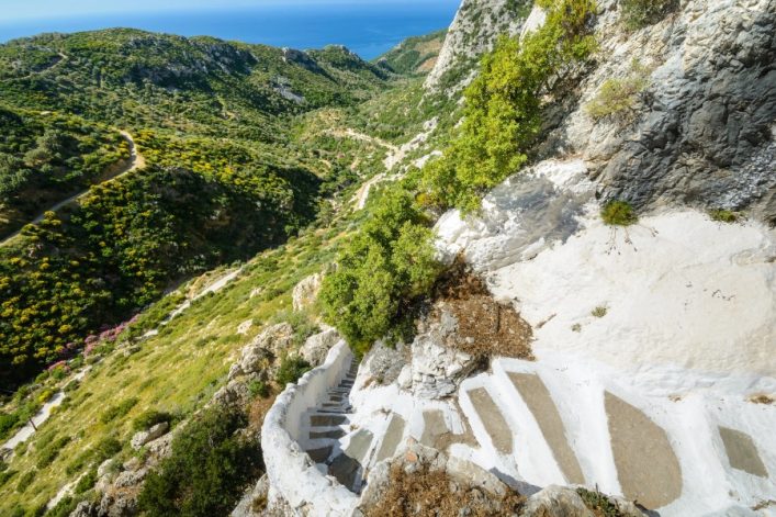 Weiße, steile Treppen führen durch grüne Hügel zur Pythagoras-Höhle auf Samos, Griechenland. Umgeben von felsigen Abhängen und üppiger Vegetation erstreckt sich in der Ferne das blaue Meer.