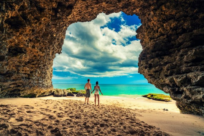 Ein Paar steht Hand in Hand am sonnigen Strand, umrahmt von Felsen, blickt aufs türkisblaue Meer, umgeben von exotischen Landschaften Balis.