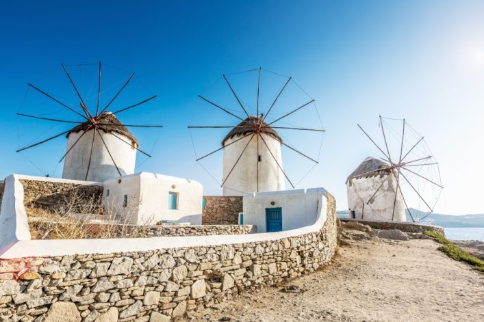 Drei traditionelle weiße Windmühlen mit kegelförmigen Dächern und großen Flügeln unter blauem Himmel, vorne eine Steinmauer auf Mykonos.