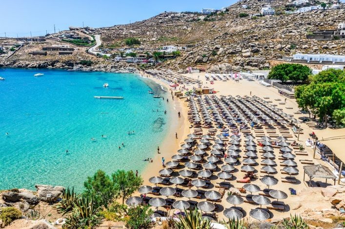Strand mit türkisblauem Wasser, vielen Badegästen und Reihen von Liegestühlen und Sonnenschirmen rechts. Im Hintergrund felsige Hügel mit vereinzelten Gebäuden, einige Bäume spenden Schatten.