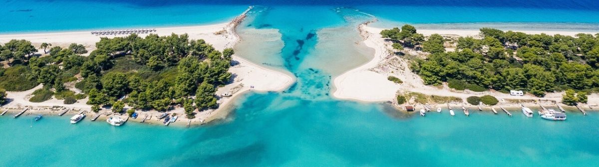 Luftaufnahme eines weißen Sandstrandes mit türkisfarbenem Wasser. Zwei bewaldete Halbinseln ragen ins Meer, rechts Boote an Stegen, links Sonnenschirme.