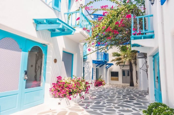 Enge Gasse mit weißen Häusern, blauen Türen und Fenstern. Pinke Bougainvillea rankt über Balkone. Der Boden besteht aus unregelmäßigen Steinplatten, im Hintergrund steht eine Palme.
