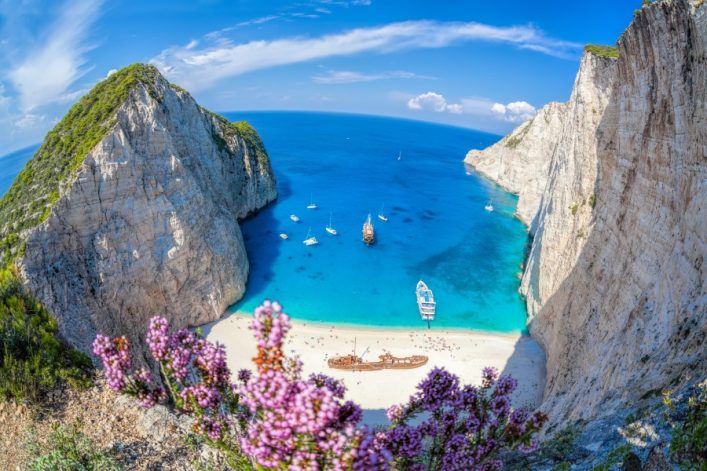 Klippen umrahmen eine türkisfarbene Bucht mit Strand, Schiff und kleinen Booten. Im Vordergrund lila Blumen, im Hintergrund blauer Himmel mit Wolken.