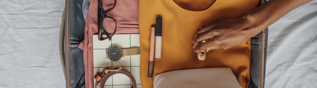 Geöffneter Koffer auf einem Bett: Brille, Uhr und Gürtel auf rosa Kleidung links; gelbes Kleid in der Mitte; daneben zwei Lippenstifte; Hand schließt Koffer; weiße Tasche unten rechts.