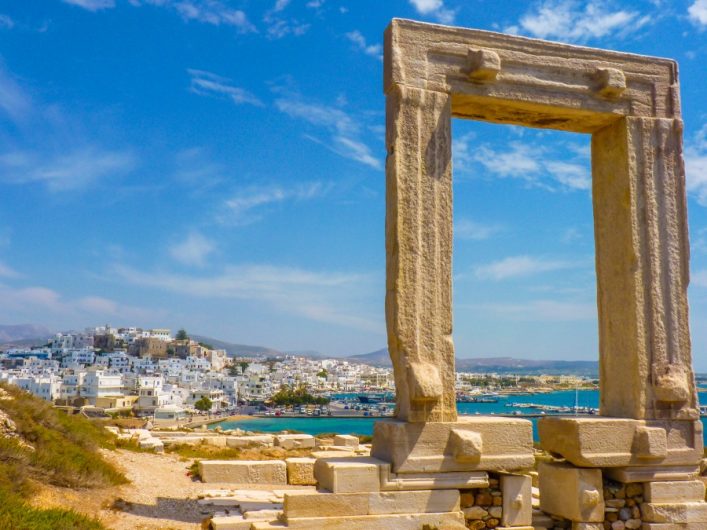 Antike Tempelruine Portara auf Naxos, Griechenland, im Vordergrund. Im Hintergrund erstreckt sich eine Stadt am tiefblauen Meer unter einem klaren, blauen Himmel.