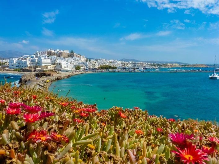 Lebhafte rote Blumen im Vordergrund, dahinter ein türkisfarbenes Meer. Am linken Ufer liegt eine strahlend weiße Stadt auf Naxos unter einem klaren, blauen Himmel. Ein Boot rechts im Wasser.