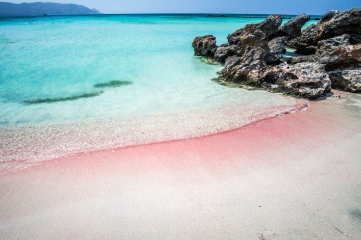 Türkisblaues Wasser trifft auf einen weißen, rosafarben schimmernden Sandstrand. Rechts ragen Felsen ins Bild, im Hintergrund sind verschwommene Berge am Horizont sichtbar.