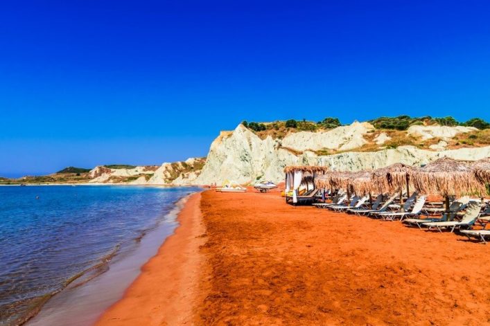 Knallorangener Sand gepaar mit den weißen Bauten im Hintergrund und davor das blaue Meer.