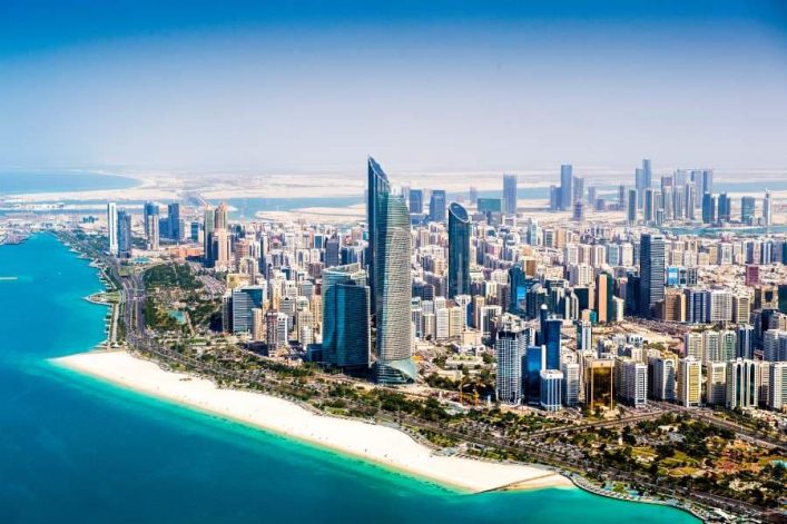 Luftaufnahme von Abu Dhabi mit Wolkenkratzern, einer küstennahen Strandlinie im Vordergrund und klarem blauem Wasser.