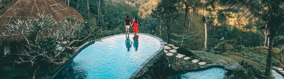 Ein Paar steht an der Spitze eines Infinity-Pools, der in einen tropischen Hang eingelassen ist, mit üppigem Grün und Palmen im Hintergrund; rechts führen Steinplatten zum Pool daneben.