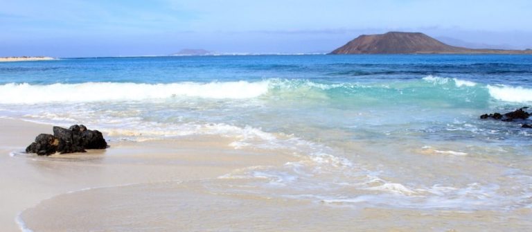 Sandstrand im Vordergrund, türkisblaues Meer mit kleinen Wellen in der Mitte, dunkle Felsen rechts, am Horizont eine braune, hügelige Insel unter blauem Himmel.