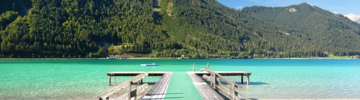 Holzsteg am Achensee führt ins klare, türkisfarbene Wasser. Berge mit dichtem Wald umgeben den See. Perfektes Urlaubsidyll am See in Österreich bei sonnigem Wetter und blauem Himmel.