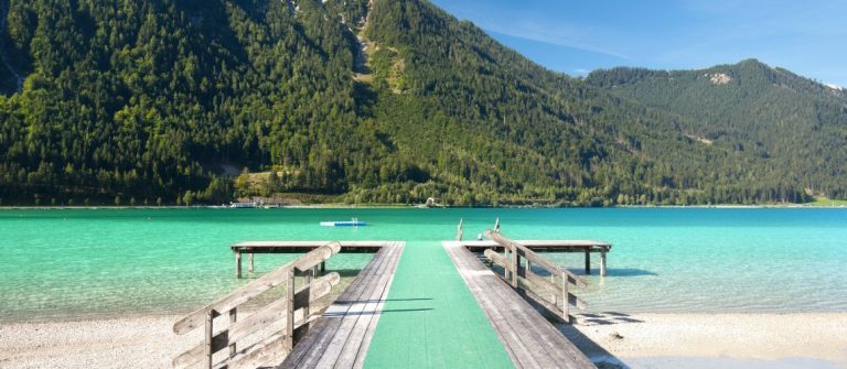 Holzsteg am Achensee führt ins klare, türkisfarbene Wasser. Berge mit dichtem Wald umgeben den See. Perfektes Urlaubsidyll am See in Österreich bei sonnigem Wetter und blauem Himmel.