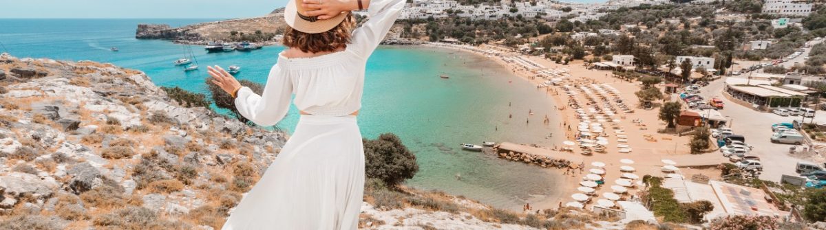 Frau in Weiß blickt auf Lindos mit seiner Akropolis auf Rhodos, umgeben von azurblauem Meer und sandigem Strand.