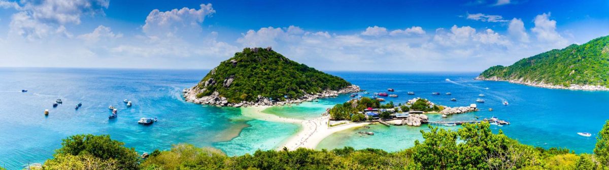 Packliste Thailand im Hinterkopf: Zwei bewaldete Inseln umgeben von türkisblauem Meer. Weißer Sandstrand verbindet Inseln, Boote ankern an Küste.