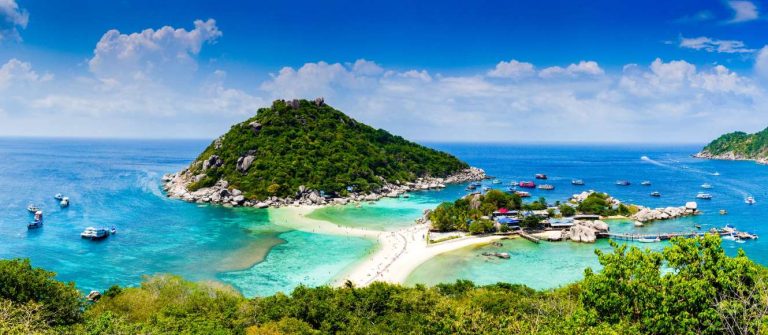 Zwei bewaldete Inseln umgeben von türkisblauem Meer. Weißer Sandstrand verbindet Inseln, Boote ankern an Küste.