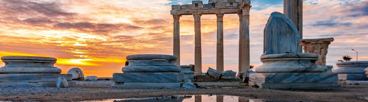 Antike Säulen des Apollo-Tempels in Side bei Sonnenuntergang, farbenfroher Himmel spiegelt sich in einer Pfütze, Ruinen umgeben die Szenerie.