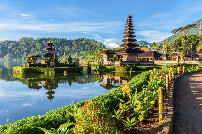 Pura Ulun Danu Bratan Tempel auf Bali spiegelt sich im ruhigen See, umrahmt von grünen Bergen und farbenfrohen Blumen im Vordergrund. Ideal für Fernreisen.