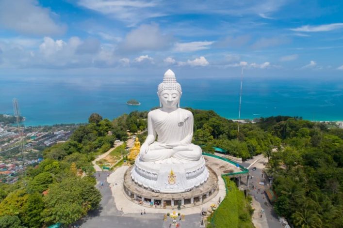 Weiße, große Buddha-Statue im Lotossitz auf einem Hügel in Phuket, umgeben von üppigem Grün und Blick auf das azurblaue Meer.
